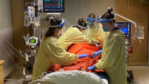 Five members of a care team dressed in personal protective equipment surround the bed of a COVID-19 patient they are caring for. 