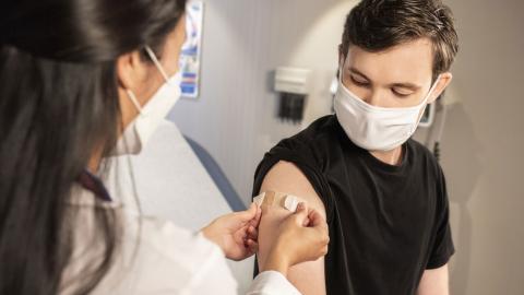 A band-aid is placed on the arm of a young man by a medical professional.