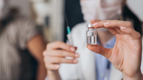 A masked health care provider holds a syringe and a vial of vaccine.