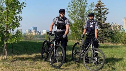 Two Protective Services Bike Officers stand with their bike in front of the Saskatoon skyline.