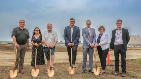 Seven people stand with grasping ceremonial golden shovels about to 'break earth' on a construction project. 