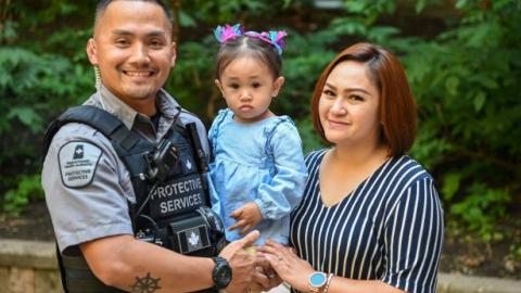 A Protective Services Officer stands, smiling, with his partner while holding his young daughter. 