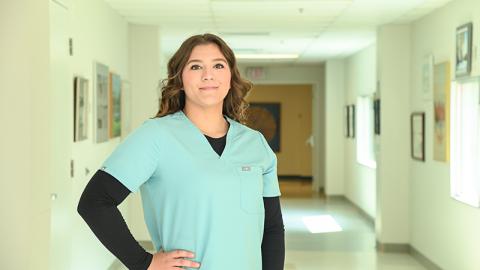 A young woman in a nurse's uniform smiles while standing in a hospital hallway. 