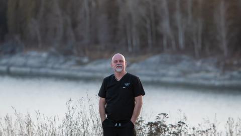 A male nurse is pictured wearing dark scrubs and standing on the shore in front of water and trees in northern Saskatchewan.