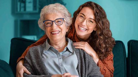 Young adult lovingly embraces senior. Both are smiling and facing the camera.