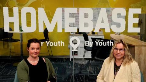 Two women sitting in front of a sign at the HOMEBASE hub in Moose Jaw.