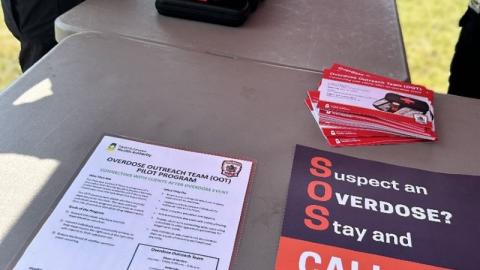 A person is standing at a table with a stack of take home naloxone kits, an informational flyer about the Saskatchewan Health Authority Overdose Outreach Team, and a poster urging anyone suspecting an overdose to call 911.