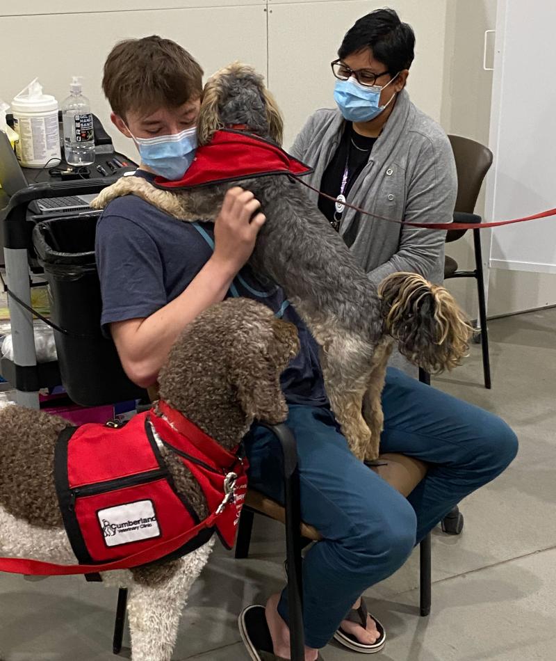 A young man sits in a chair with a dog on his lap and a dog by his side, while an SHA employee in the background gets ready to give him his vaccine. 