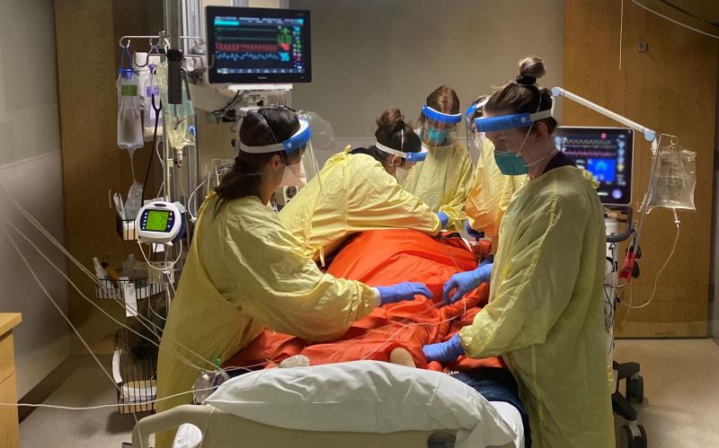 Five members of a care team dressed in personal protective equipment surround the bed of a COVID-19 patient they are caring for. 