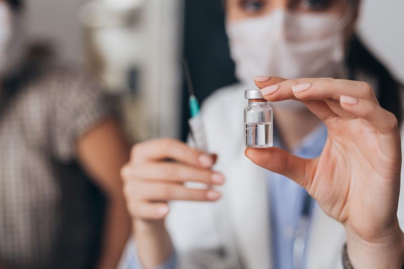 A masked health care provider holds a syringe and a vial of vaccine.