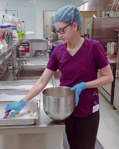 Reanna Hawkins, a Cook, holds a mixing bowl in her left hand while scooping batter onto a cooking pan with her right hand.