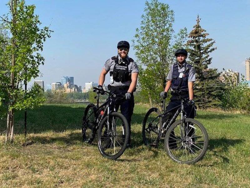 Two Protective Services Bike Officers stand with their bike in front of the Saskatoon skyline.