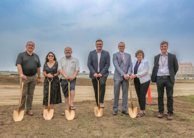 Seven people stand with grasping ceremonial golden shovels about to 'break earth' on a construction project. 