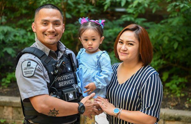 A Protective Services Officer stands, smiling, with his partner while holding his young daughter. 