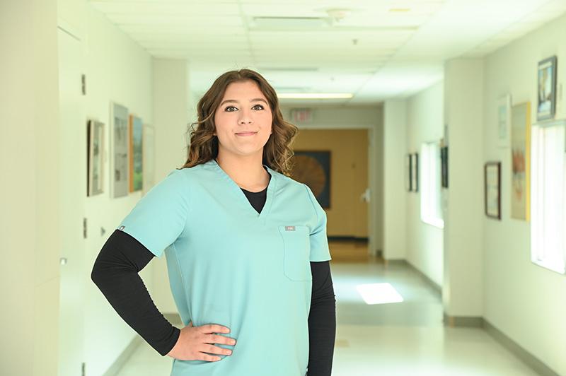 A young woman in a nurse's uniform smiles while standing in a hospital hallway. 