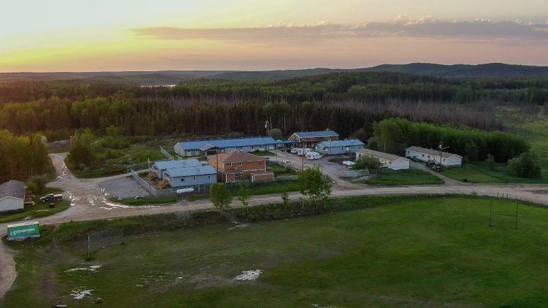 An aerial view of the Sandy Bay Health Centre shows the facility on a road with a thick forest of trees and a beautiful sunset in the background.