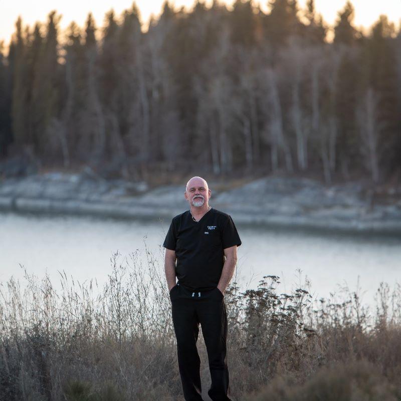 A male nurse is pictured wearing dark scrubs and standing on the shore in front of water and trees in northern Saskatchewan.