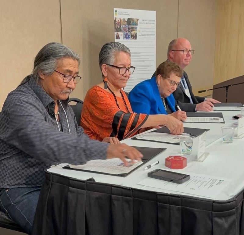 Knowledge Keepers Daniel Alphonse and Judy Pelly, SHA Board Chair Arlene Wiks, and SHA CEO Andrew Will sit at a table signing the SHA's reaffirmation of its commitment to Truth and Reconciliation.