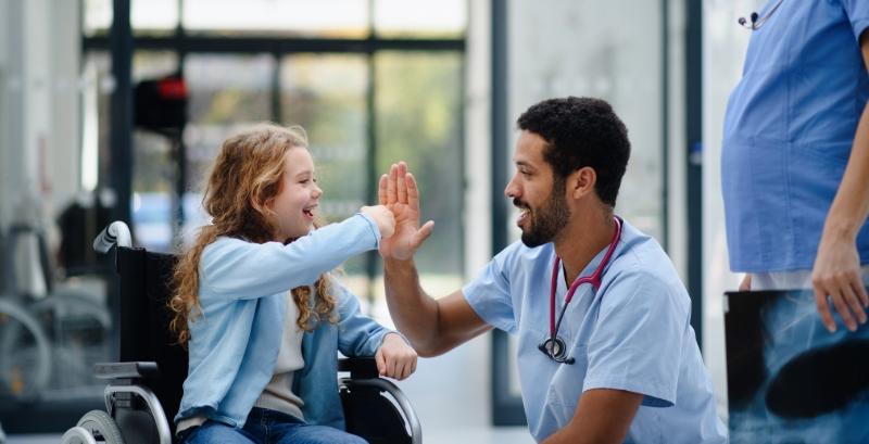 A health-care worker wearing scrubs high fives a girl in a wheelchair.