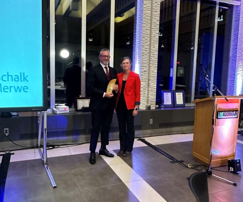 Dr. Schalk Van der Merwe and President of Saskatchewan College of Family Physicians, Dr. Darcie McGonigle pose together, at an award ceremony. Both are dressed formally and Dr. Van der Merwe holds an award for the 2024 Family Physician of the Year – Saskatchewan.