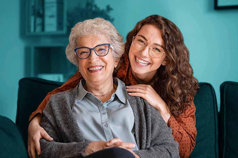 Young adult lovingly embraces senior. Both are smiling and facing the camera.