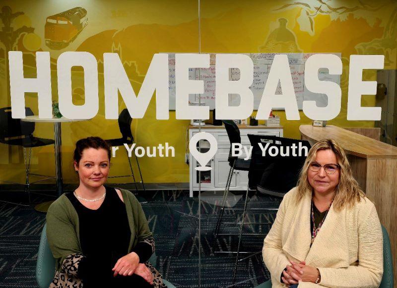 Two women sitting in front of a sign at the HOMEBASE hub in Moose Jaw.