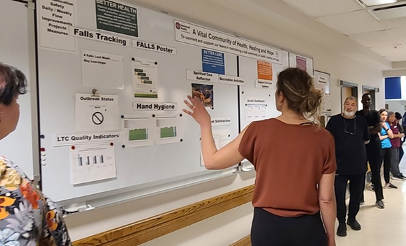 A lady in an orange shirt stands in front of a whiteboard. The whiteboard has several forms and posters on it, illustrating an example of visual management.